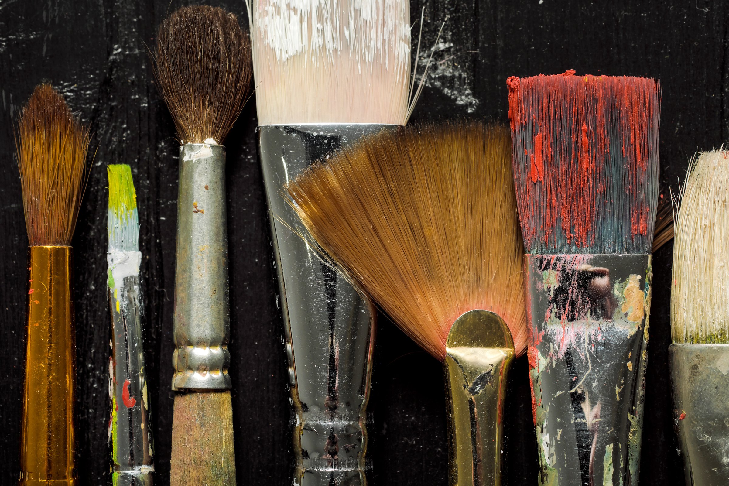 Artist's tools, brushes in paint are in a row on a black wooden background.